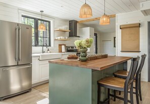 A modern kitchen with a stainless steel refrigerator, wooden island, hanging pendant lights, and a view of the outdoors through large windows. - Darling 4 bed 2 bath cottage on Lake Muskoka, minutes from Bala (Parry Sound Mid-Shore (Bala))