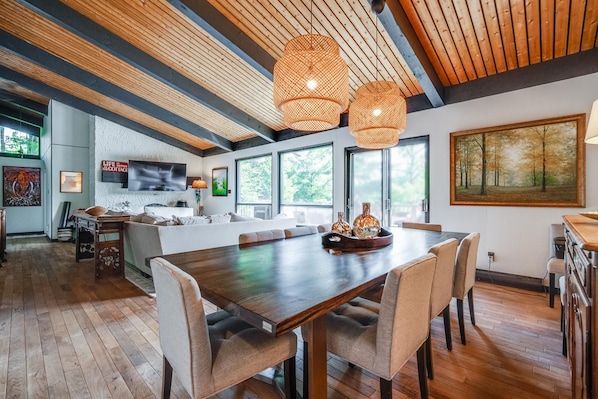 Een ruime woonkamer met een grote houten eettafel, comfortabele zitplaatsen en een aan de muur bevestigde televisie.