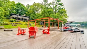 Eine Holzterrasse mit roten Adirondack-Stühlen, einem überdachten Sitzbereich und einem am Wasser liegenden Boot.