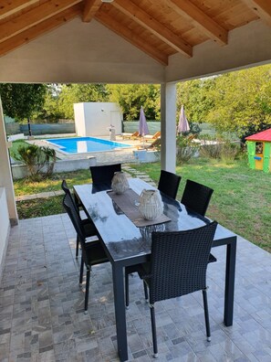 Eine überdachte Terrasse mit einem rechteckigen Tisch und schwarzen Korbstühlen, mit Blick auf ein Schwimmbecken und eine Rasenfläche mit einem kleinen Spielhäuschen.
