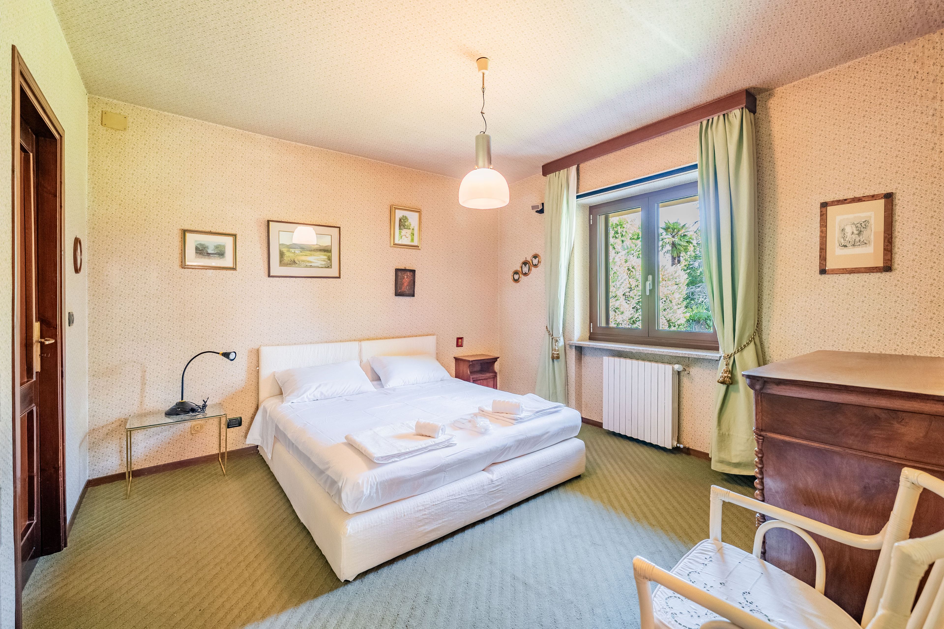 Et hotelværelse med seng, natbord, lampe, vindue med gardiner og billeder på væggen.