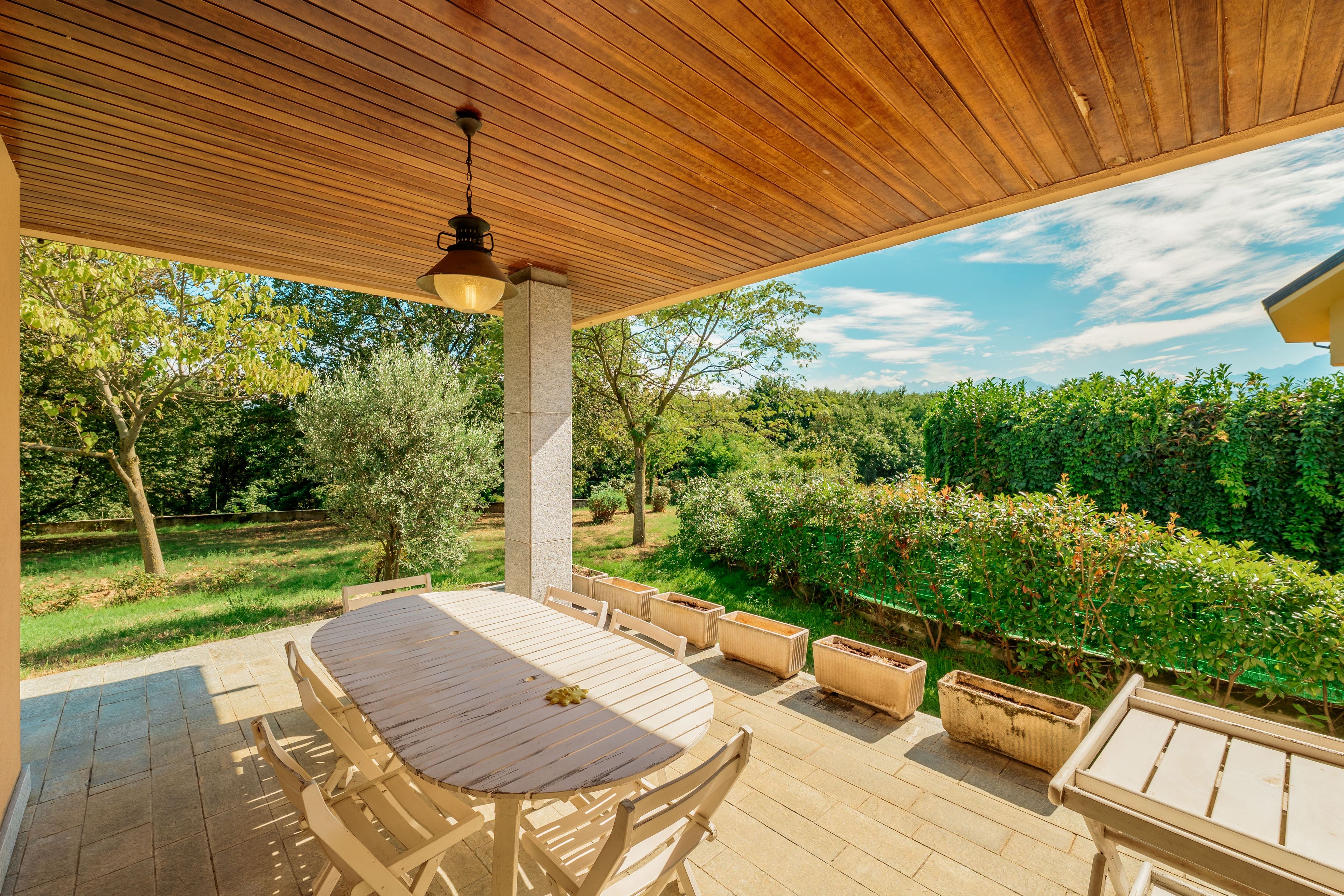 An outdoor patio with a wooden table and chairs, surrounded by greenery and a clear sky.