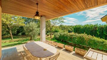 An outdoor patio with a wooden table and chairs, surrounded by greenery and a clear sky.