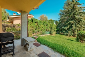 Un pati amb una barbacoa, seients exteriors i vistes a un jardí i arbres ben cuidats.