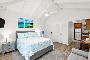 A bedroom with a bed, bedside tables, a water dispenser, a ceiling fan, and a view of a kitchen area.