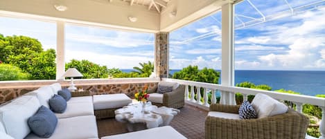 A covered outdoor seating area with a view of the ocean, furnished with sofas and a coffee table.