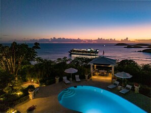A pool area with lounge chairs and umbrellas, a gazebo, and a view of the ocean and a distant city skyline at dusk. - Bellevue Estate- 6BR New remodel w/Restoration Hardware & Tennis (St. Thomas)