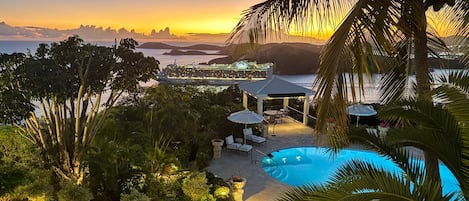 A resort with a swimming pool, lounge chairs, and a view of the sea at sunset.