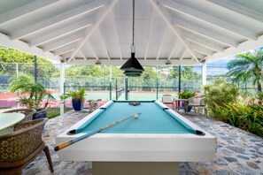 A pool table with a hanging light fixture, surrounded by wicker chairs and potted plants, under a white roofed area with a view of a tennis court. - 2BR New remodel w/Restoration Hardware & Tennis (Charlotte Amalie East)