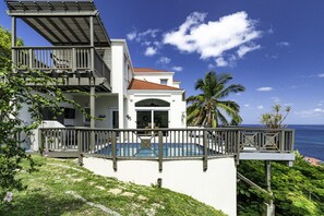 Ein zweistöckiges Haus mit Balkon und Blick auf den Ozean, einem Schwimmbad und üppiger Bepflanzung.