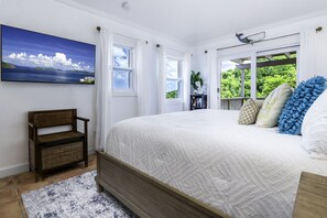 Ein Schlafzimmer mit einem großen Bett, einem Korbsessel, einem Gemälde und Blick auf Grün durch das Fenster.