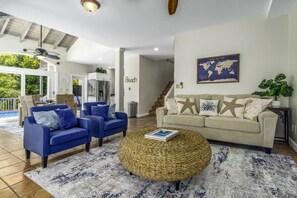 Ein Wohnzimmer mit einem blauen Sofa, einem beigen Sofa und einem runden Kaffeetisch aus Korbmöbeln. An der Wand hängt eine Weltkarte und eine Treppe führt in einen anderen Raum.