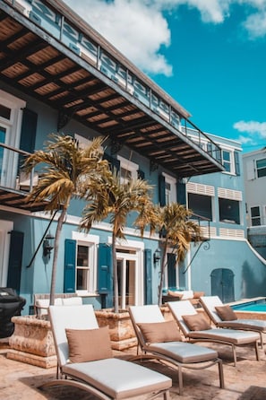 A poolside area with lounge chairs and palm trees, a staircase, and a building with blue shutters. - Lissette Manor - 5BR historical estate next to The Pink Palm Hotel downtown! (St. Thomas)