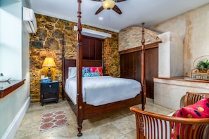 Uma cama de dossel com um painel de parede de pedra, um criado-mudo de madeira com abajur, e uma cesta de vime sobre uma mesa consola.