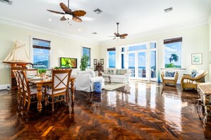 A spacious living room with a wooden herringbone floor, a dining area with a wooden table and chairs, a white sofa, wicker chairs, and a large window with a view of the ocean. - Lissette Manor - 5BR historical estate next to The Pink Palm Hotel downtown! (St. Thomas)