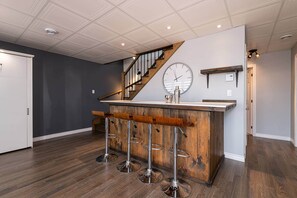 A bar area with a wooden counter and four stools, a clock on the wall, and a staircase in the background. - Le Feu Follet: Riverview Home (La Malbaie)
