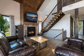 A spacious living room with a fireplace, leather sofas, a wooden coffee table, and a staircase with a railing. - Le Feu Follet: Riverview Home (La Malbaie)