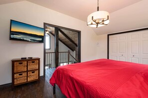 A bedroom with a red bedspread, a wooden chest of drawers, a painting, and a staircase leading to another room. - Le Feu Follet: Riverview Home (La Malbaie)