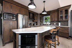 A modern kitchen with dark wood cabinets, stainless steel appliances, a large island, and pendant lighting. - Le Feu Follet: Riverview Home (La Malbaie)