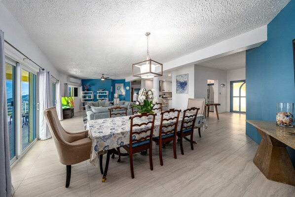A spacious dining area with a long table and chairs, a ceiling light, and a view of the outdoors through large windows. - 4BR - centrally located & mins. from Magen`s Bay! (St. Thomas)