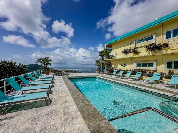 Un espace piscine avec des chaises longues, une vue dégagée sur l’océan et un bâtiment doté de balcons.