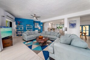 A spacious living room with a large sectional sofa, a coffee table, and a TV. There are shelves with decorative items and a ceiling fan. - 4BR - centrally located & mins. from Magen`s Bay! (St. Thomas)