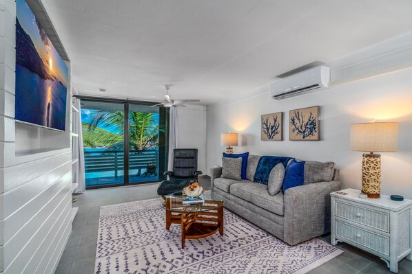 A living room with a sofa, coffee table, and a TV on the wall. There is a window with a view of palm trees. - Stunning Beachfront Condo & Pool (St. Thomas)
