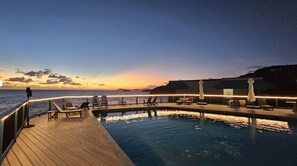 Ein Poolbereich mit Liegestühlen und Sonnenschirmen mit Blick auf das Meer bei Sonnenuntergang.