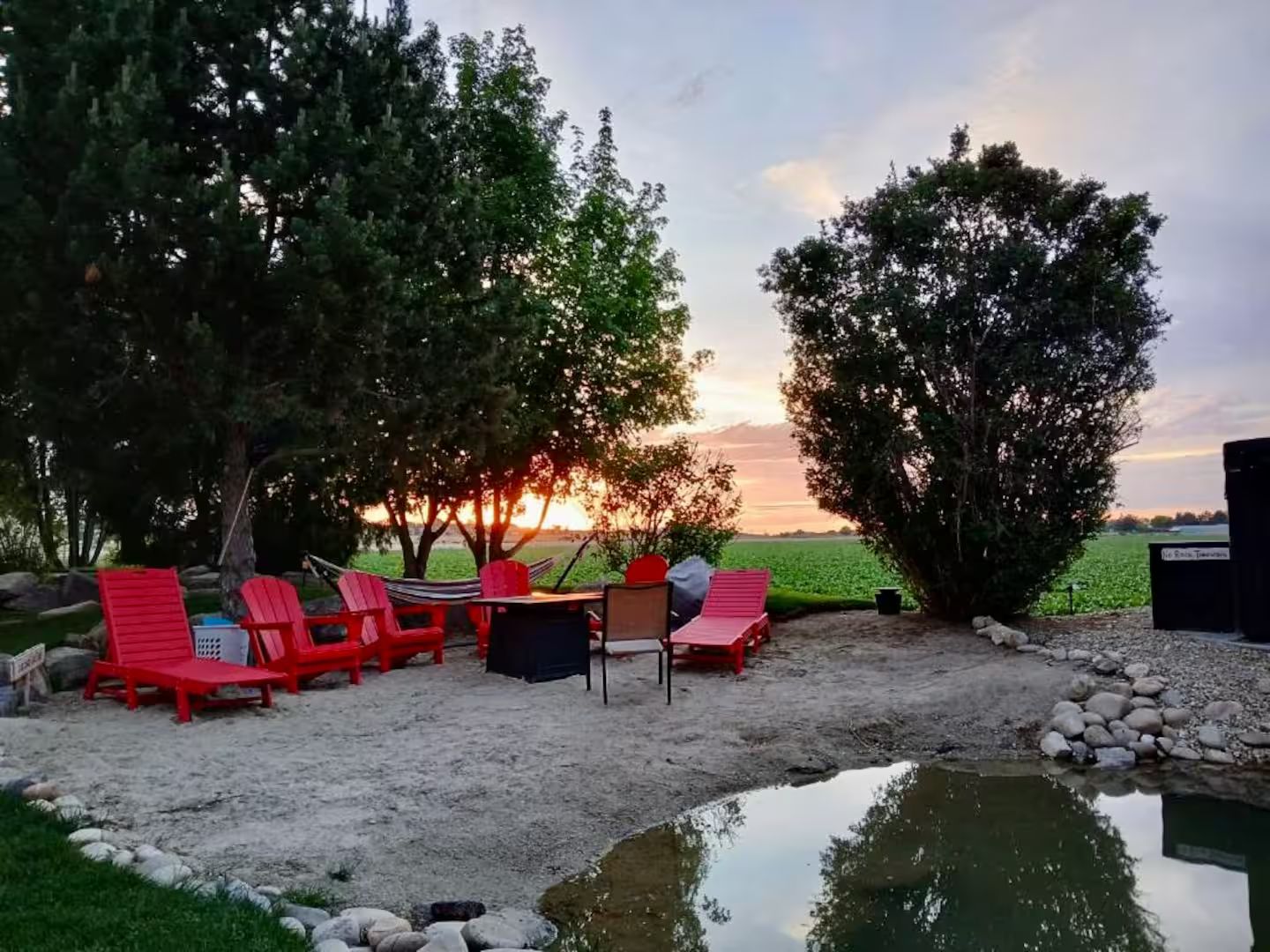 A serene outdoor seating area with red chairs and a table, surrounded by trees and a view of the sunset.
