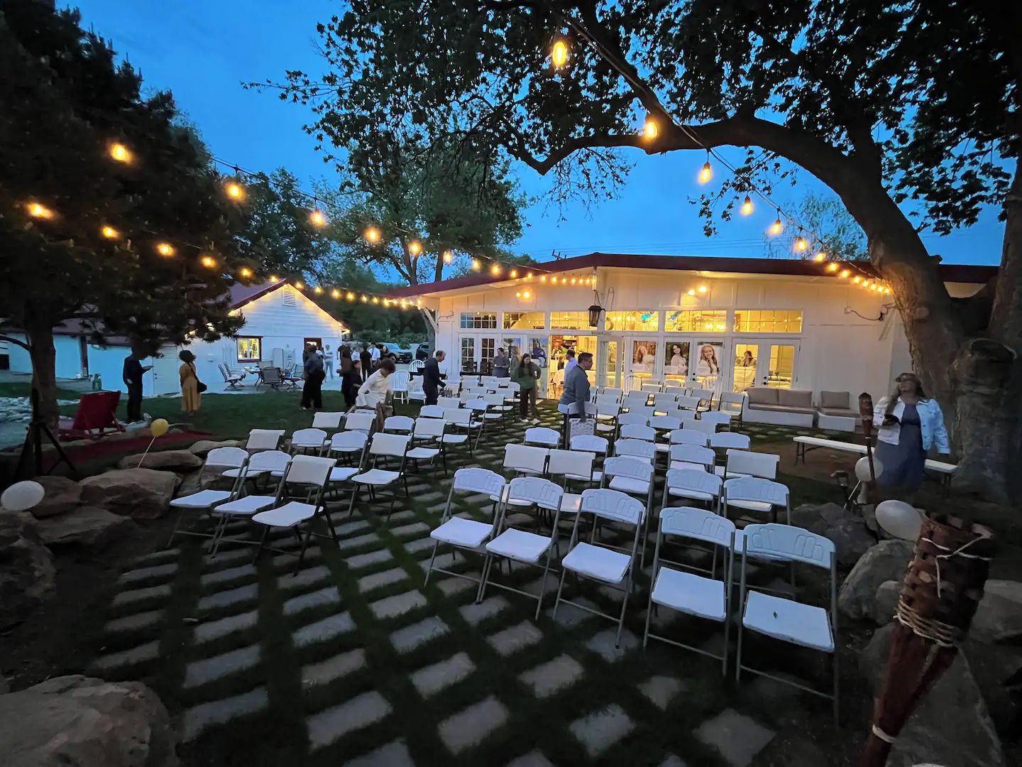 Een buitenzitplaats met rijen stoelen, ingericht voor een evenement, onder een grote boom met lichtslingers.