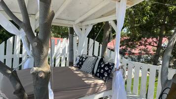 A covered outdoor seating area with a bench and cushions, surrounded by white picket fencing and trees.