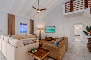 A spacious living room with a high ceiling, a ceiling fan, a large flat-screen TV, a sofa, a wicker sofa, a coffee table, and a potted plant. - 5BR Villa w/pool in peaceful residential neighborhood (Charlotte Amalie West)