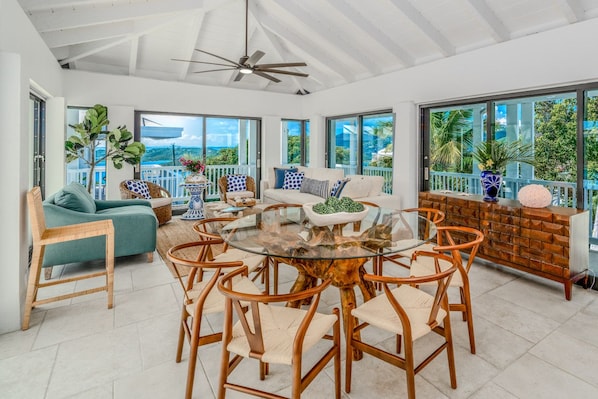 A spacious living area with a glass-top coffee table surrounded by wicker chairs, a sofa, and a wooden cabinet. Large windows offer views of the outdoors. - Sunset Sanctuary: Gorgeous 5br w/huge pool/deck & lots of space! (St. Thomas)
