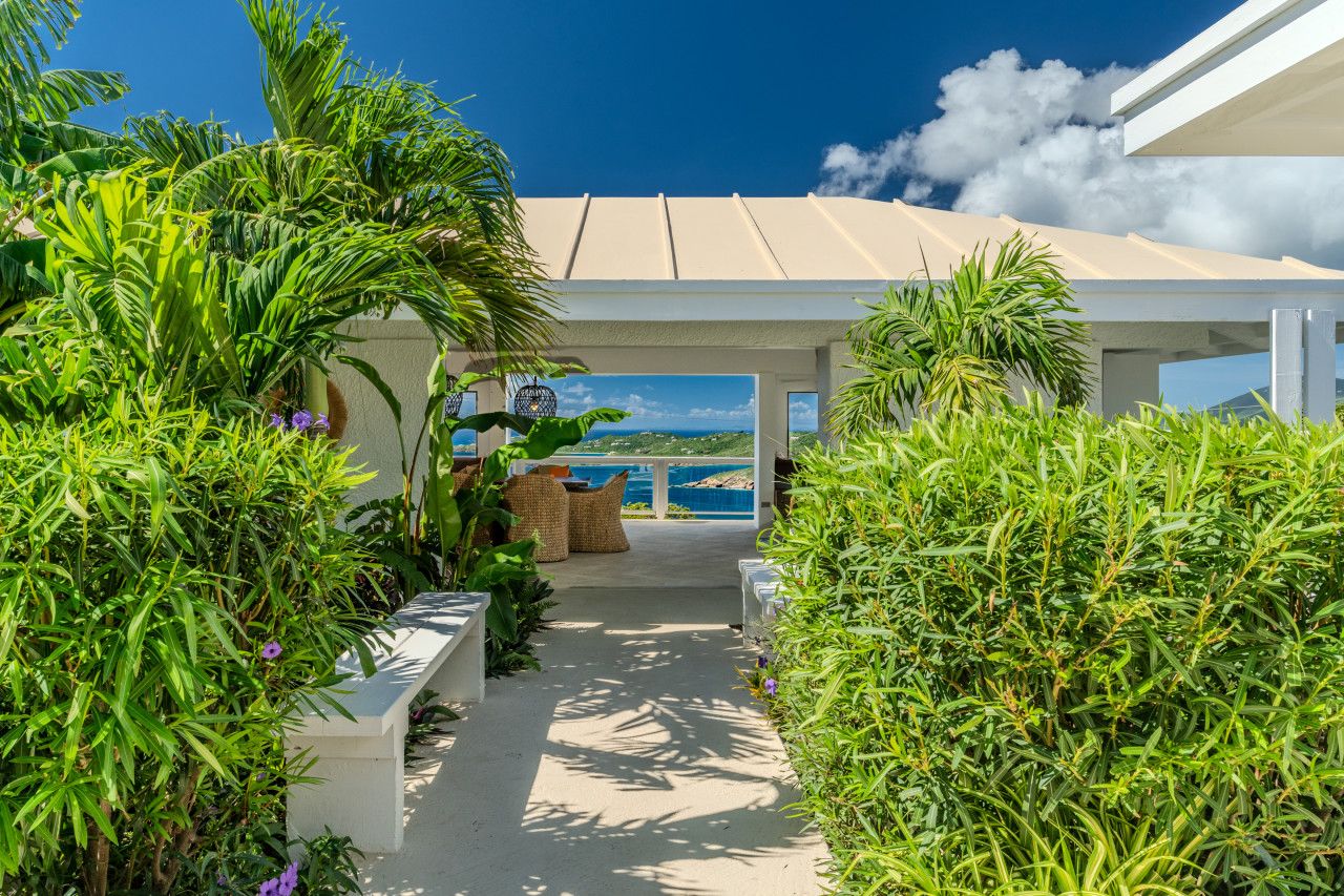 A covered outdoor area with a view of the sea, surrounded by lush greenery and a pathway leading to a seating area.