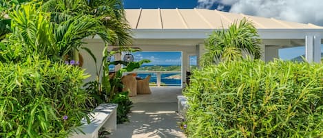 A covered outdoor area with a view of the sea, surrounded by lush greenery and a pathway leading to a seating area.
