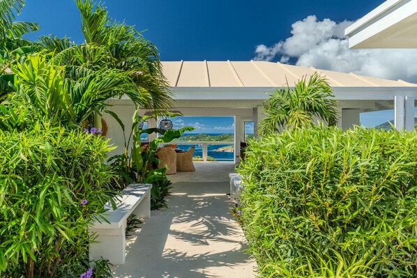 A covered outdoor area with a view of the sea, surrounded by lush greenery and a pathway leading to a seating area. - Sunset Sanctuary: Gorgeous 5br w/huge pool/deck & lots of space! (St. Thomas)