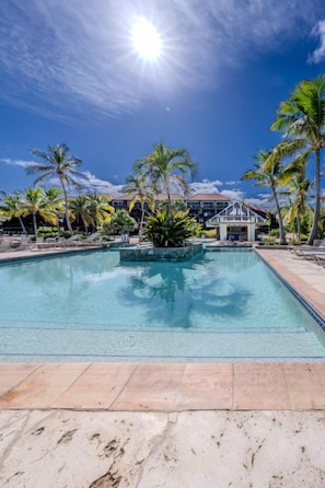 A swimming pool with lounge chairs and palm trees, surrounded by a building with a tiled roof. - 1Br condo in Sapphire Beach Resort - pool & beach! Sleeps 4 (St. Thomas)