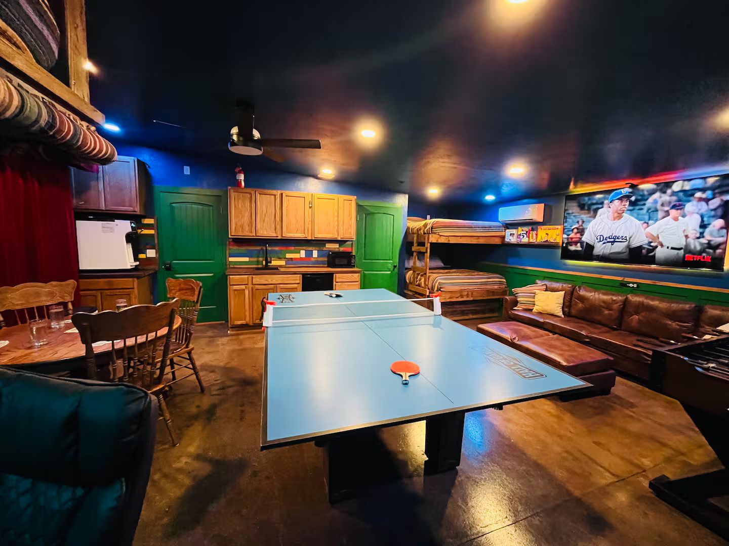 A room with a ping pong table, seating area, kitchen, and a television displaying a sports event.