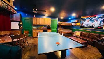 A room with a ping pong table, seating area, kitchen, and a television displaying a sports event.
