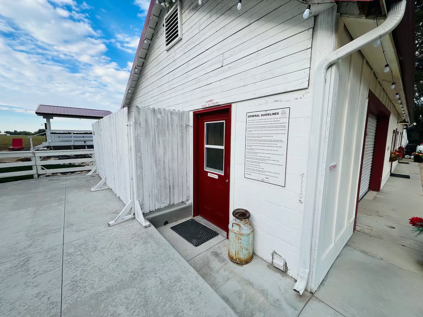 A white building with a red door, a sign with general guidelines, and a metal container on the ground.
