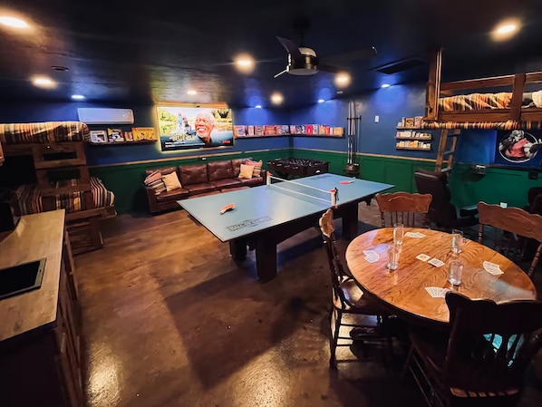 A room with a ping pong table, a sofa, a round table with playing cards, and a television displaying a movie.