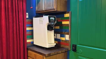 A small kitchenette with a refrigerator, a microwave, and a coffee machine. There is a wooden cabinet above the counter and a green door to the right.