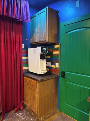 A small kitchenette with a refrigerator, a microwave, and a coffee machine. There is a wooden cabinet above the counter and a green door to the right.