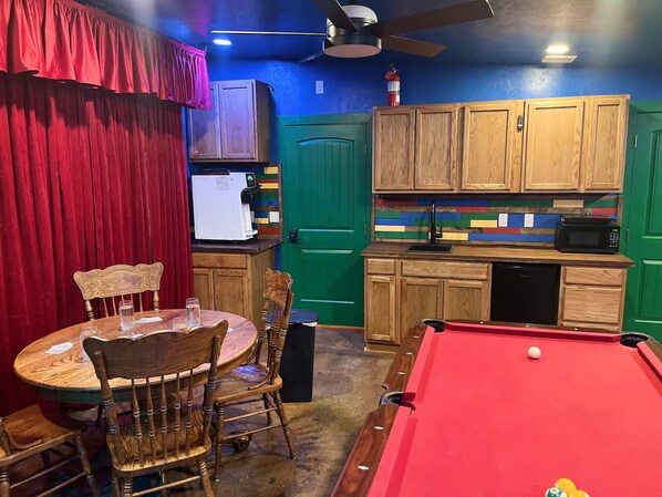 A room with a pool table, a dining table with chairs, a microwave, and a refrigerator.