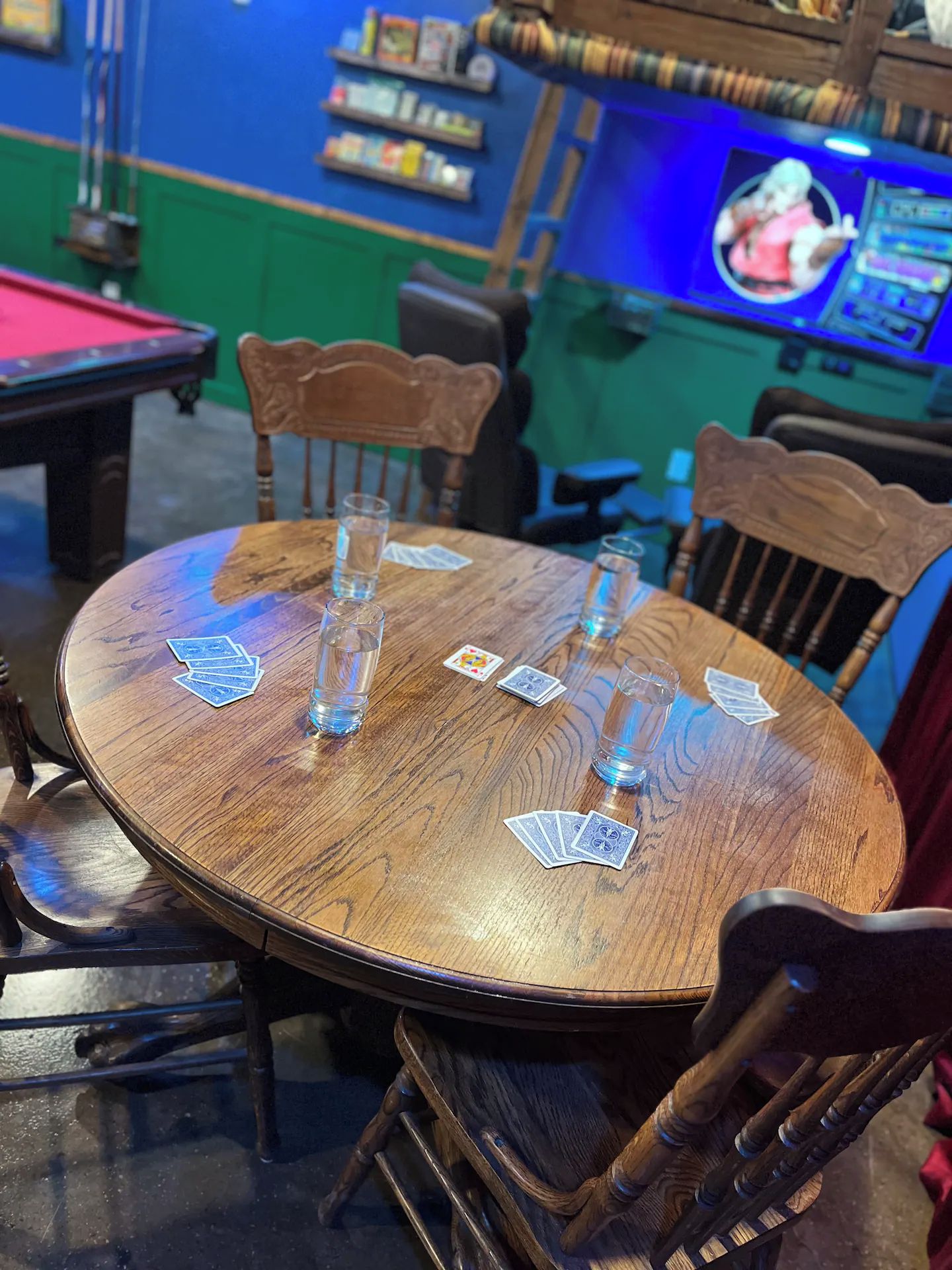 Una mesa de madera con cartas y vasos de chupito, rodeada de sillas, en una habitación con una mesa de billar y una pantalla de televisión.