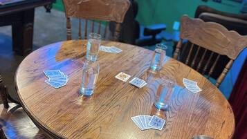 Una mesa de madera con cartas y vasos de chupito, rodeada de sillas, en una habitación con una mesa de billar y una pantalla de televisión.