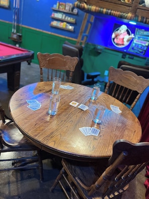 A wooden table with playing cards and shot glasses, surrounded by chairs, in a room with a pool table and a television screen.