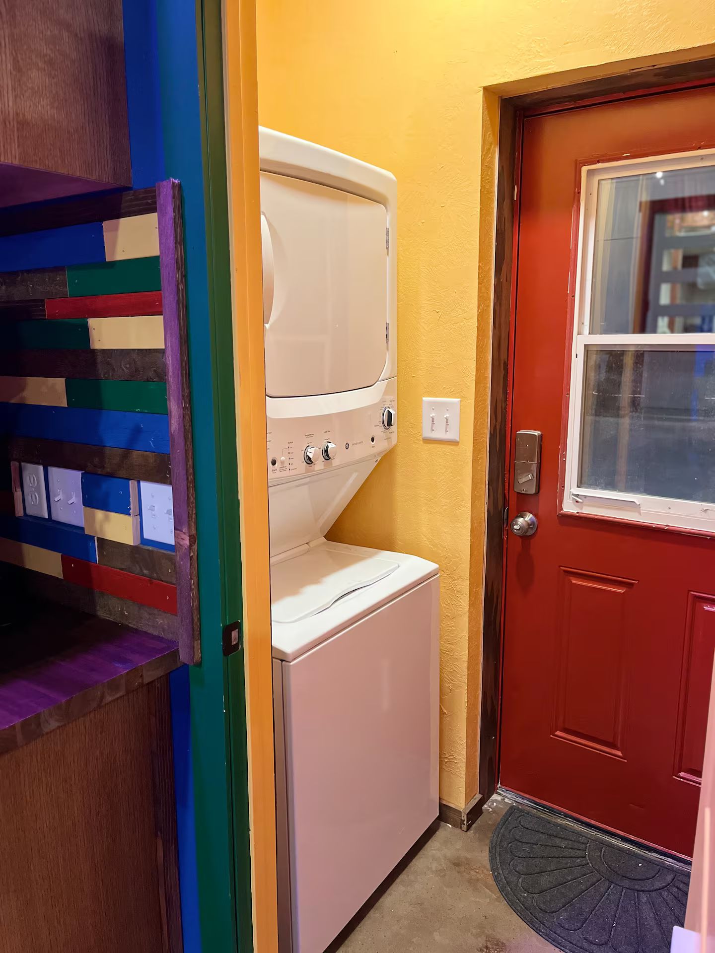 Un espace buanderie équipé d’un lave-linge et d’un sèche-linge, avec une porte rouge et un mur aux couleurs vives.