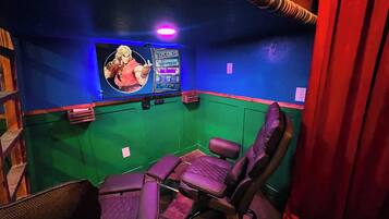 A room with a green wall, a blue accent wall, and a large screen displaying a character. There are two purple gaming chairs and a wooden shelf.