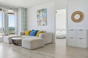 A modern living room with a sofa, ottoman, and a round coffee table. There is a painting on the wall and a round woven wall decor. - Marina Retreat by IIM (Estepona)